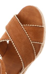 Bruine suède sandaal met kruisbandjes, versierd met gouden kralendetails en een getextureerde wigzool. Close-upweergave van het schoenontwerp.