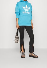 Modrá mikina s bílým logem Adidas, černé legíny s bočními rozparky, sandály s květinovým vzorem a taška s zebra vzorem.