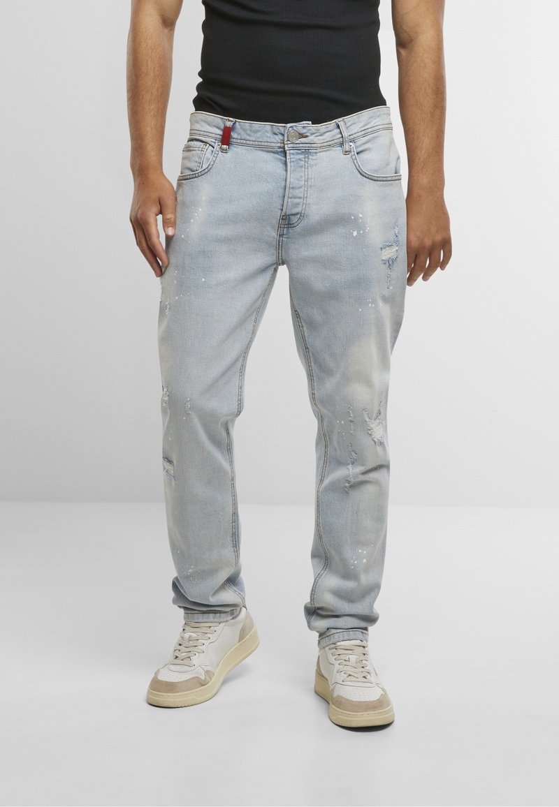 Homme portant un jean bleu clair usé avec des taches de peinture, un débardeur noir et des baskets beiges, debout devant un fond uni.