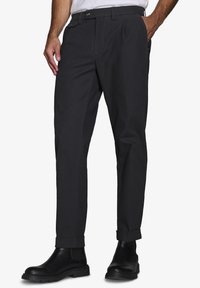 Pantaloni neri con taglio dritto, vita plissettata, orli con risvolto, tasche laterali e chiusura con bottone. Indossati con stivaletti neri alla caviglia.