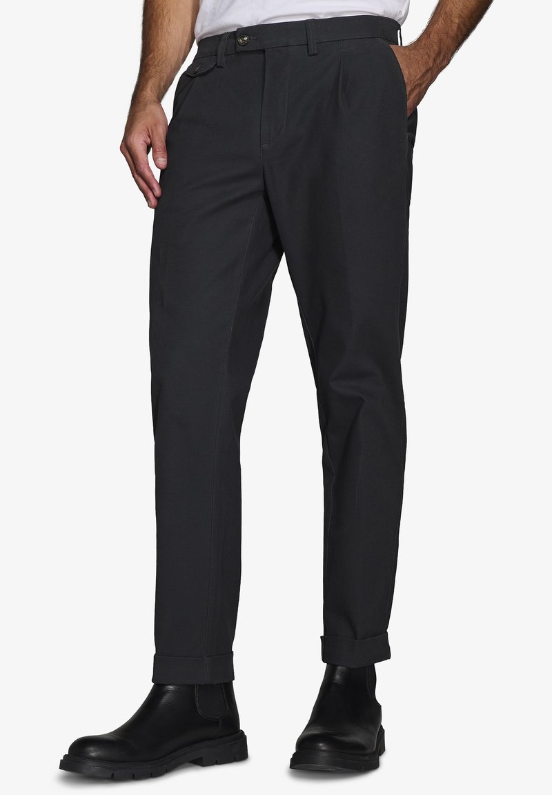 Pantalones negros con corte recto, pliegues en la parte delantera, dobladillos con puños, bolsillos laterales y cierre de botón. Llevados con botas negras de tobillo.