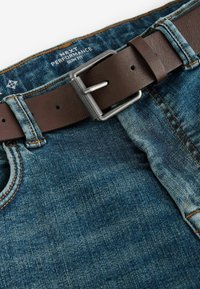 Curea din piele maro prinsă pe blugi de un albastru închis, având cusături deschise și un design slim fit. Eticheta scrie „NEXT PERFORMANCE.”