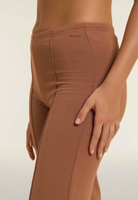 Leggings marroni realizzati in materiale morbido e elasticizzato con una superficie liscia. Presentano un'etichetta con logo discreto e cuciture verticali per un aspetto sartoriale.