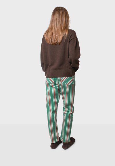 Pullover marrone oversize con orlo a costine, abbinato a pantaloni verdi con righe verticali rosse e bianche, indossato con pantofole marroni scure.