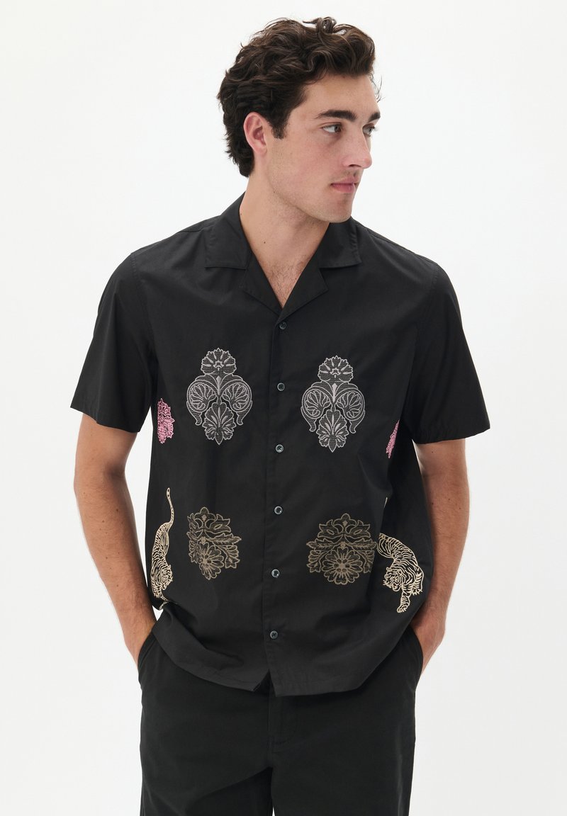 Camisa negra de manga corta con bordados de patrones florales y de animales en rosa, dorado y blanco. Presenta un cierre de botones en la parte delantera y un cuello.