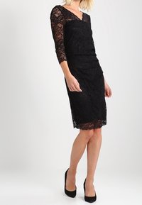 Robe en dentelle noire avec des manches trois quarts, silhouette ajustée, décolleté en V et ourlet festonné. Associée à des chaussures à talons hauts noires.