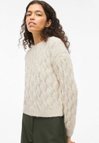 Pull beige en maille torsadée avec un col rond et des manches longues, présentant un motif texturé ainsi que des poignets et un ourlet côtelés.