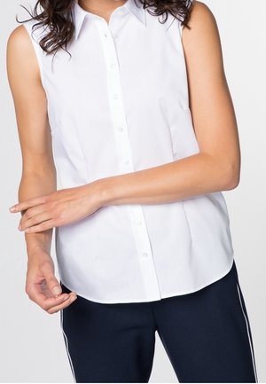 Donna che indossa una camicia bianca senza maniche con colletto e bottoni e pantaloni blu navy con strisce bianche laterali, braccia incrociate in vita.