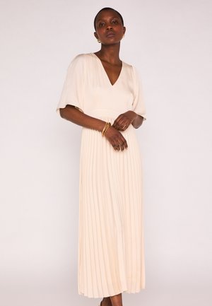 Femme aux cheveux courts portant une robe midi plissée crème, des boucles d'oreilles créoles dorées et des bracelets dorés, sur un fond clair uni.