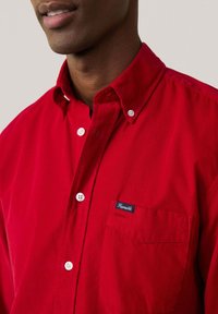 Camisa roja de botones confeccionada en tela con un acabado texturizado. Presenta un bolsillo en el pecho y un pequeño parche con el logo. Los botones blancos contrastan con el color.