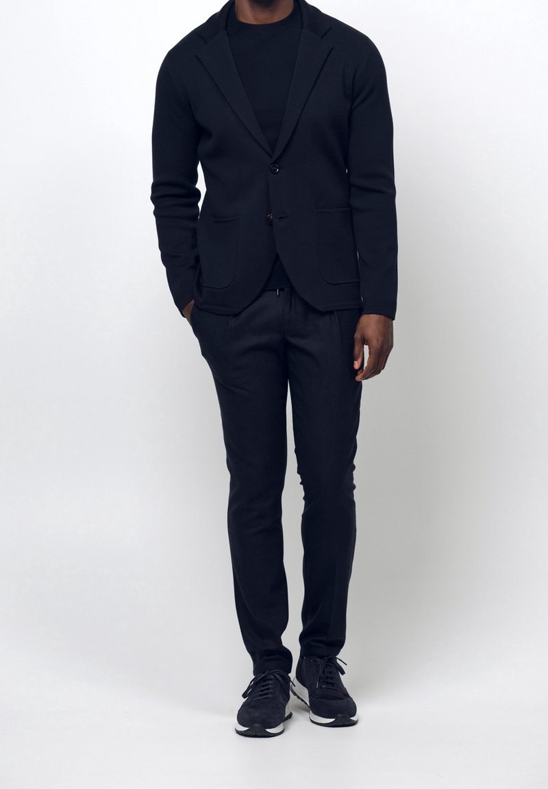Homme portant un blazer noir, une chemise noire, un pantalon noir et des baskets noires, debout devant un fond blanc uni.