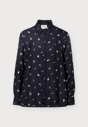 Personne portant une chemise noire à manches longues avec des motifs nautiques blancs et un pantalon beige à jambes larges, debout devant un fond uni.