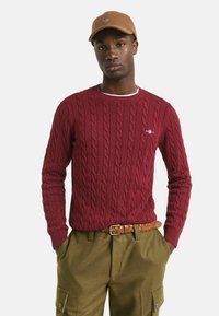 Junger Mann in braunem Hut, bordeauxrotem Strickpullover, olivgrünen Cargo-Hosen und mit einem geflochtenen braunen Gürtel, der mit Händen in den Taschen steht.