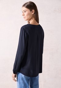Blusa navy a maniche lunghe con maniche testurizzate, scollo rotondo, dettaglio della piega posteriore e una vestibilità rilassata, abbinata a pantaloni di denim chiaro.