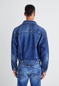 Denim jacket in dark blue with long sleeves, a pointed collar, and button closures. Features pockets and a slightly distressed texture.