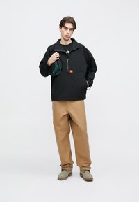 Μαύρο πουλόβερ με μισό φερμουάρ και κόκκινο λογότυπο, συνδυασμένο με ανοιχτό καφέ παντελόνι και πράσινα αξεσουάρ, με άνετη εφαρμογή και τσέπες.