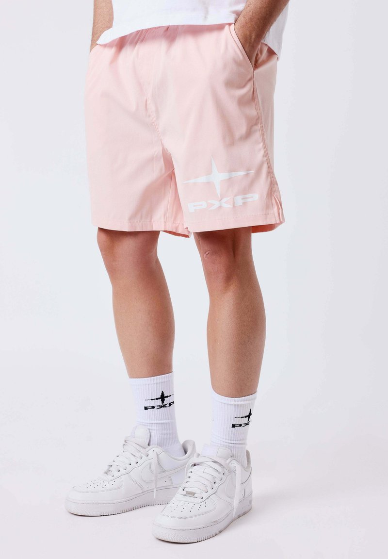 Personne portant un short rose clair avec le logo blanc "PXP", des chaussettes blanches montantes avec le même logo, et des baskets blanches à lacets. Mains dans les poches.