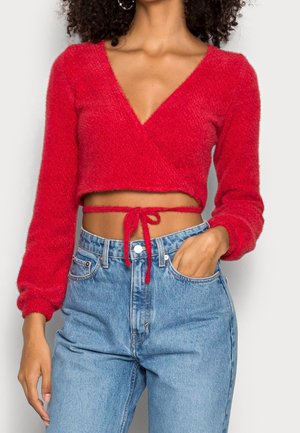 Femme portant un crop top rouge duveteux à manches longues, noué à la taille, associé à un jean bleu clair taille haute, main dans la poche.