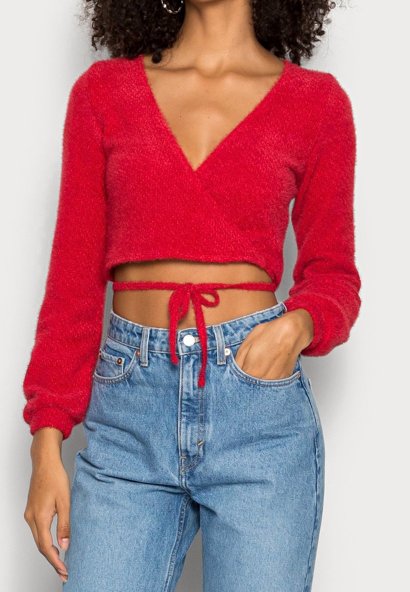 Femme portant un crop top rouge duveteux à manches longues, noué à la taille, associé à un jean bleu clair taille haute, main dans la poche.