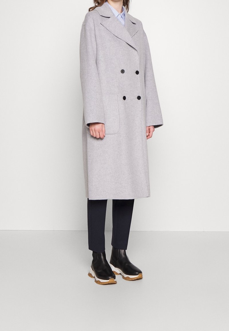 Personne portant un long manteau croisé gris clair, un pantalon noir et des bottes noires à semelles blanches, sur un fond clair uni.