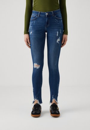 Skinny blue denim jeans with distressed areas, frayed hems, and a fitted design. Worn with black sneakers featuring a gum sole.