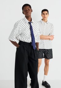 Dva modely nosia oblečenie s bodkami: jeden v bielom tričku s čiernymi bodkami, širokých čiernych nohaviciach a modrom kravate; druhý v tričku s krátkym rukávom a bodkovaných šortkách.