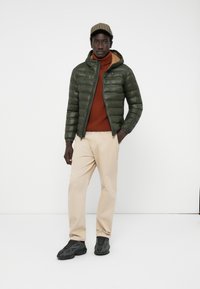 Dark green puffer jacket with a hood, brown turtleneck, beige trousers, and black sneakers. Features quilted texture and zip closure.