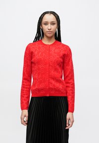 Rood vest met bloemmotief, ronde hals, knopen aan de voorkant en lange mouwen. Draagt over een geplooide zwarte rok. Gladde textuur.