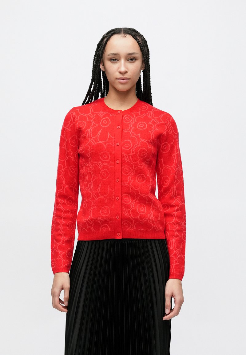 Rood vest met bloemmotief, ronde hals, knopen aan de voorkant en lange mouwen. Draagt over een geplooide zwarte rok. Gladde textuur.