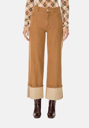 Donna che indossa pantaloni marroni a vita alta e gamba larga con grandi risvolti beige, abbinati a una maglia a maniche lunghe a quadri e stivaletti marrone scuro.