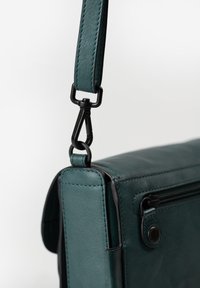 Türkisfarbenes Leder-Umhängetasche mit schwarzem Metallzubehör, ausgestattet mit einem abnehmbaren Tragegurt, einem Reißverschlussfach und einem kantigen Design. Glatte Textur, minimalistische Details.