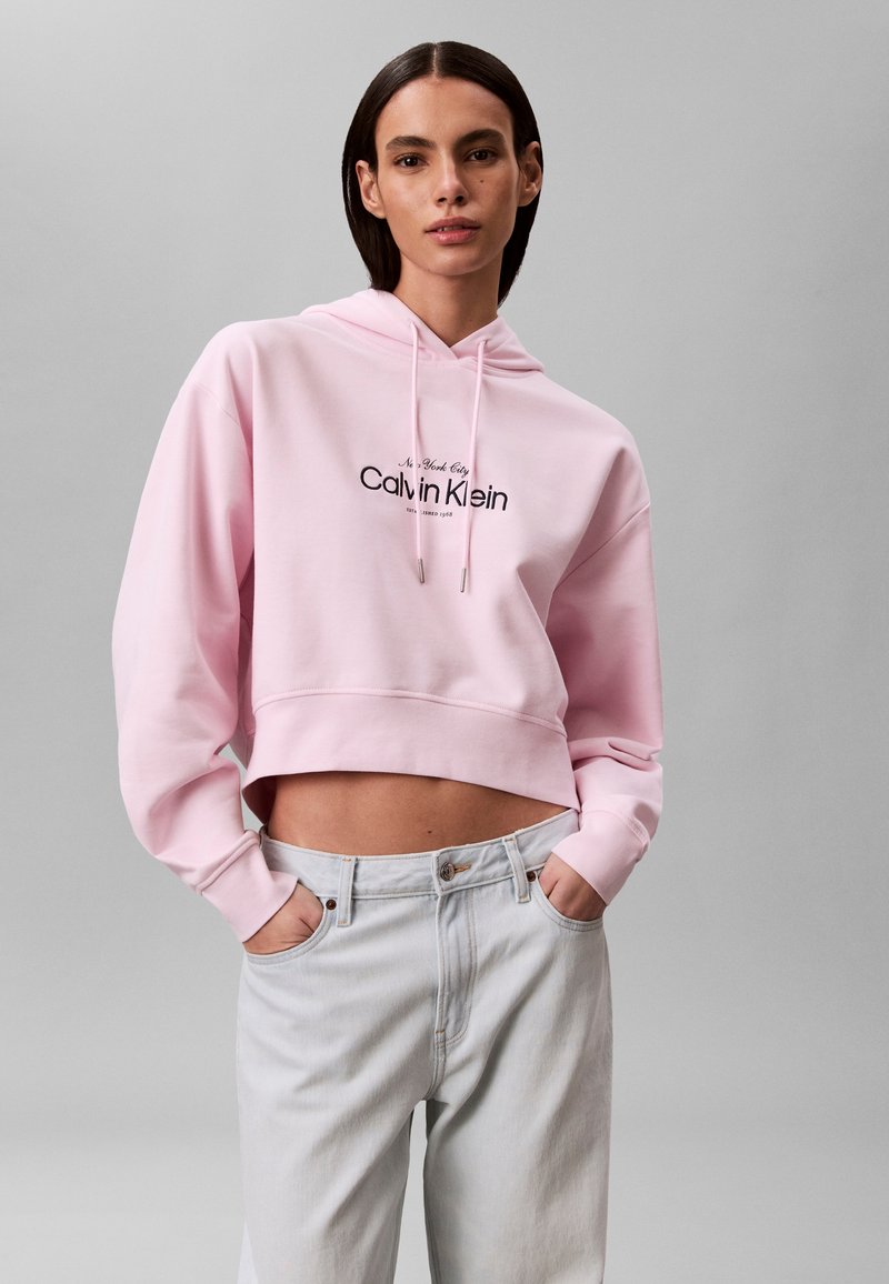 Νέα γυναίκα που φοράει ένα ροζ κοντό hoodie Calvin Klein και ανοιχτό μπλε τζιν, στέκεται με τα χέρια στις τσέπες μπροστά σε ένα γκρι φόντο.