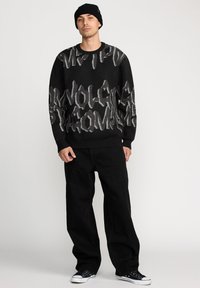 Pull en tricot noir avec des lettres abstraites grises, associé à un pantalon large noir et des baskets basses noires. Le mannequin porte un bonnet noir.