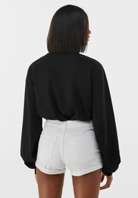 Donna con capelli lunghi e lisci che indossa una maglietta nera a maniche lunghe e pantaloni corti bianchi con orlo arrotolato, girata di spalle su uno sfondo neutro.