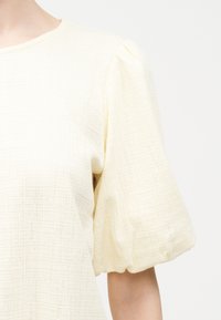 Εκρού υφασμάτινο τοπ με υφή, με κοντά μανίκια φουσκωτά και στρογγυλή λαιμόκοψη σε ένα άτομο.