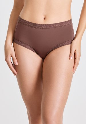 Frau in braunen, hochgeschnittenen Slips mit floralem Spitzenbesatz an der Taille und den Beinöffnungen, die entspannt mit den Armen an den Seiten steht.