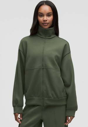 Donna con felpa verde oliva a collo alto con zip e pantaloni abbinati, in piedi davanti a uno sfondo chiaro e uniforme.