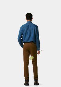 Denimhemd in Mittelblau kombiniert mit braunen Slim-Fit-Hosen. Das Modell hält eine weiße Blume, trägt dunkle Stiefel und ist von hinten zu sehen.