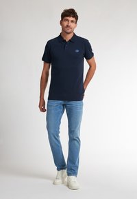 Námořnické polokošile s modrým logem, krátkými rukávy a klasickým límcem. Kombinováno se světle modrými džíny a bílými teniskami.