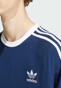 T-shirt blu navy con colletto bianco e tre righe orizzontali sulle maniche, caratterizzato da un logo ricamato bianco sul lato sinistro del petto.