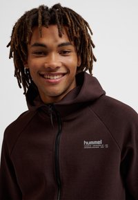 Młody mężczyzna z dredami, uśmiechający się, ubrany w ciemnobrązową bluzę z kapturem na zamek błyskawiczny z logo "hummel" na piersi, na jednolitym tle.