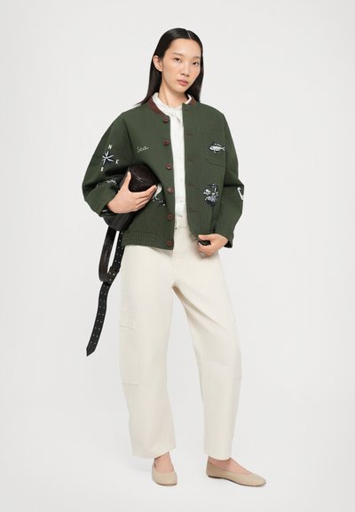 Groene geborduurde bomberjack met ronde kraag, knoopsluiting, gecombineerd met crèmekleurige wijde broek en beige flats, met een donkere tas in de hand.