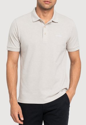 Mann trägt ein beiges Polohemd mit kurzen Ärmeln, zwei Knöpfen und kleinem Logo auf der linken Brustseite, kombiniert mit schwarzen Hosen.