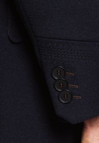 Morski wełniany żakiet z rękawem, na którym znajdują się trzy czarne guziki z brązowymi przeszyciami i subtelną teksturą materiału.