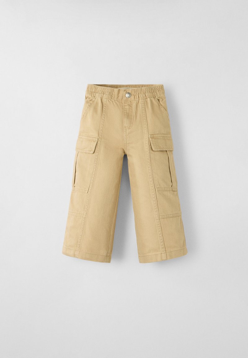 Calças cargo bege com uma cintura elástica, apresentando dois bolsos laterais e dois bolsos frontais com aba. Feitas de um tecido de algodão durável.