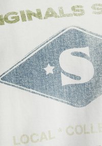 Un material textil alb prezintă un grafic de denim albastru în formă de diamant, evidențiind un „S” proeminent și o stea, cu text verde deasupra și dedesubt.