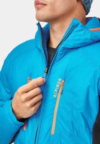 Niebieska ocieplana kurtka z kapturem, z pomarańczowymi akcentami, zamykanymi kieszeniami i lekkim materiałem. Regulowane sznurki w kapturze.