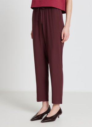 Donna che indossa pantaloni larghi rosso scuro con coulisse in vita e décolleté scuri con punta affilata e tacco basso, in piedi su un pavimento chiaro.