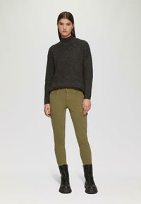 Donker geribbelde trui met hoge hals, gecombineerd met olijfgroene cargobroek met zijzakken. Zwarte veterschoenen maken de outfit compleet.