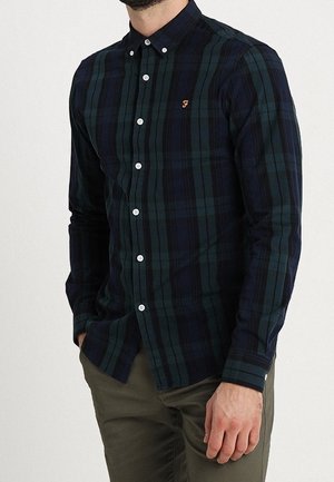 Mężczyzna ubrany w ciemnozieloną i granatową koszulę w kratę zapinaną na białe guziki oraz oliwkowe spodnie, ręce w kieszeniach.